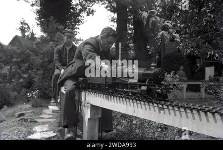 1950s, historical, outside in a garden, model railway enthusiasts at a miniature garden steam railway, at the Field End Railway Co, England, UK.  On a raised track sitting on concrete posts, two men riding on the tiny steam trains, one being a scaled model of the GWR 'County of Oxford' (1023 ) steam locomotive, the original of which started service in 1947. Stock Photo