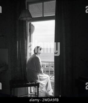 1950s, historical, early morning and a lady in a nightdress and quilted bed top sitting on a wooden slated luggage bench inside a hotel bedroom by its balcony looking out at the sea, England, UK. Stock Photo