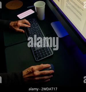 A professional's desk at night, with the focus on hands typing and a wireless mouse, highlighted by the screen's soft glow. Stock Photo