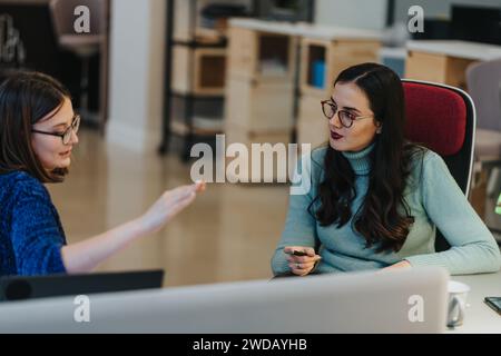 A focused view of two women conversing in a bustling co working environment, with other creative professionals collaborating in the background. Stock Photo