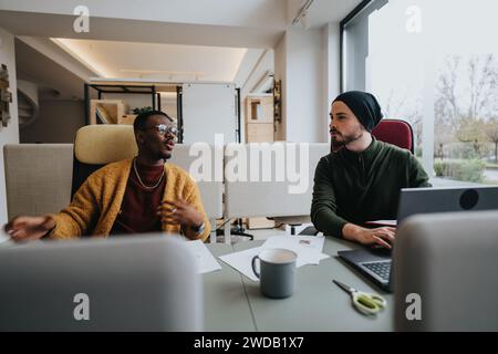 Two diverse male colleagues in a modern office engage in a productive discussion. Casual work attire reflects a relaxed corporate culture. Stock Photo