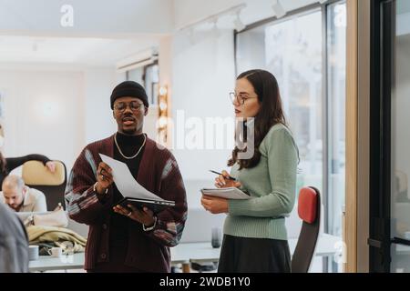 Two people, a man and woman, stand in a well-lit office space, exchanging ideas with paper notes in hand, as colleagues collaborate in the background. Stock Photo