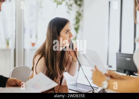 Focused female professional engaged in a business call while colleague hands over document in bright office Stock Photo