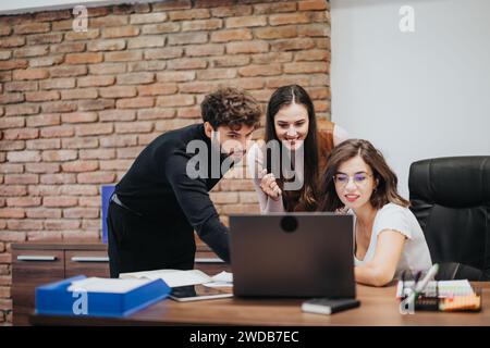 Three professionals collaborating at a laptop in a modern office with a brick wall backdrop, displaying teamwork and engagement. Stock Photo