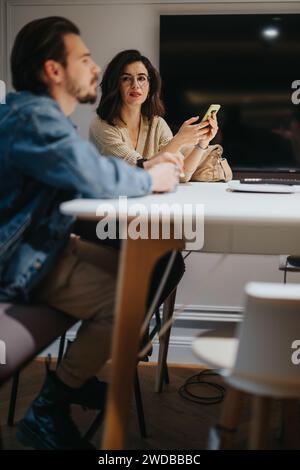 A woman with glasses holding a smart phone looks contemplative while sitting opposite a man in a casual contemporary home setting. Stock Photo