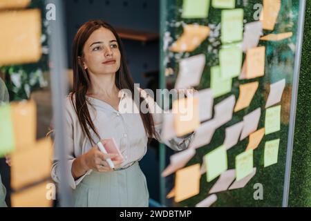 A diverse group in collaborative office. Glass wall covered in sticky notes with stats, ideas, and project costs. Woman writing, calculating profit. Stock Photo