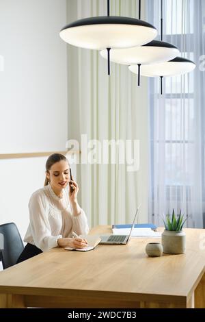 Young woman in office making notes while talking on smartphone Stock Photo