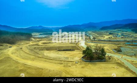 Aerial view of construction site with small cluster of trees fenced off for protection and rolling hills under blue sky in background, South Korea Stock Photo