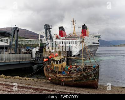 An old fishing boat moored at the harbour in front of a large ship with cloudy sky and mountains in the background, ship cutters ashore in Ullapool Stock Photo