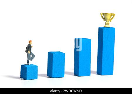 Miniature people toy figure photography. A boy pupil student running above increasing wooden ladder with trophy. Isolated on a white background. Image Stock Photo