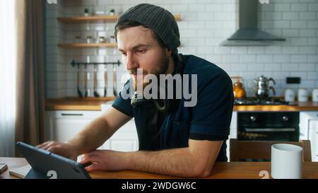 Focused businessman with beanie working at home in the kitchen working with his tablet computer, taking notes, doing research. Busy freelancer working Stock Photo