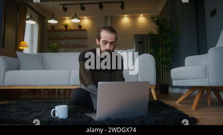Bored young bearded man sitting on the floor, using laptop computer does remote work Stock Photo