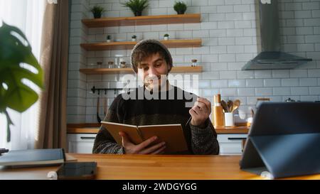 Smiling student with beanie sitting in modern kitchen at home looking at the notes in notebook he is holding in his hand, reading, thinking and changi Stock Photo