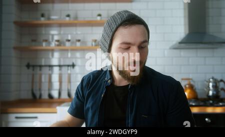 Portrait of young businessman with beanie sitting in modern kitchen at home, talking in video conference call. Confident male have video conference, r Stock Photo