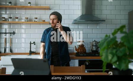 Handsome young adult man talking on the phone in modern kitchen at home. Pleasant conversation of a man with his family and friends Stock Photo