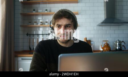 Handsome student with beanie sitting in kitchen at home wear wireless headset having video conference calling on laptop computer talk by webcam, learn Stock Photo