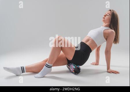 Caucasian woman doing roller exercise for myofascial release on white background. Self-massage of thighs.  Stock Photo