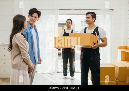 Professional movers aid couple indoors unloading boxes from truck into the home. Service guarantees Stock Photo