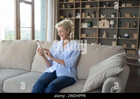 Mature woman enjoys chatting on-line sit on sofa with smartphone Stock Photo