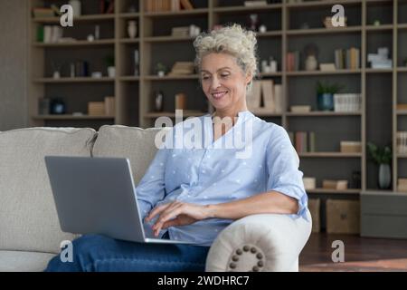 Smiling mature woman sit on couch with modern laptop Stock Photo