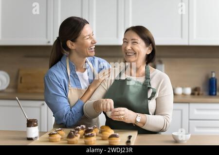 Joyful adult daughter woman helping senior mom to decorate cupcakes Stock Photo