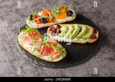 Delicious bruschetta with arugula, ham, vegetables, cream cheese close-up on a slate board on the table. Horizontal Stock Photo