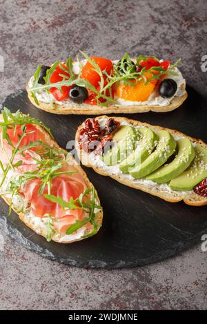Fresh open sandwiches with arugula, jamon, sun-dried tomatoes, avocado, cream cheese, olives and roasted peppers close-up on a slate board on the tabl Stock Photo
