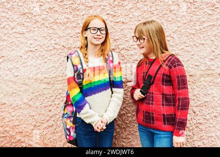 Group of two adorable kid girls posing outdoors against pink wall, wearing glasses, school backpacks and bright colorful pullovers, back to school con Stock Photo