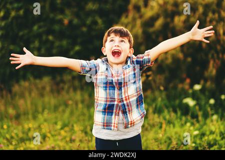Happy kid boy of 6 year old having fun outdoors wearing blue plaid shirt, arms wide open Stock Photo