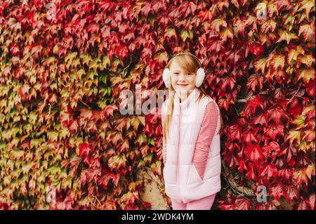 Outdoor portrait of pretty little girl posing against red wall covered with red ivy leaves, wearing pink warm vest and earmuffs Stock Photo