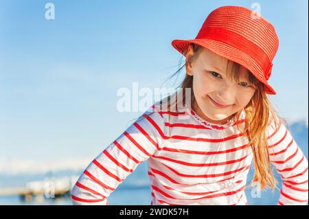Outdoor portrait of adorable little kid girl against sunny blue sky, wearing red hat and stripe shirt Stock Photo