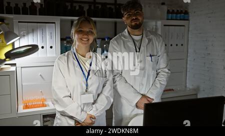 Man and woman in white lab coats are coworkers in a laboratory with computer equipment, looking professional and collaborative. Stock Photo