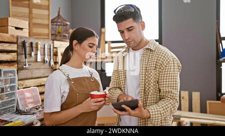 Man and woman carpenters collaborate in a woodshop, discussing over a digital tablet while holding coffee. Stock Photo