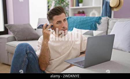 Focused young hispanic man using laptop while sitting on floor at home, intently listening to voice message on smartphone Stock Photo