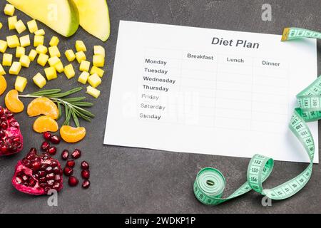 Sheet of paper with diet plan for week. Measuring tape on table. Mango, pomegranate, kiwi fruit on black background. Flat lay. Healthy Diet Plan. Stock Photo