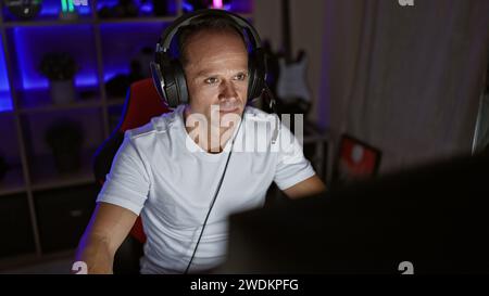 Handsome middle age man streaming video game at night, sitting seriously concentrated at his cyber gaming room, fully equipped with futuristic technol Stock Photo