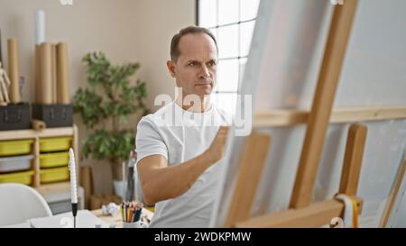 Concentrated middle age man artist passionately drawing in studio, immersed in creativity with brush, canvas and palette Stock Photo