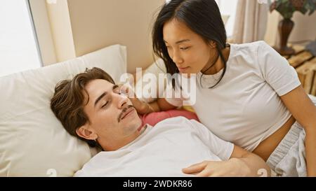 Interracial couple sharing a tender moment in a cozy bedroom setting, with a woman looking lovingly at a man lying down Stock Photo