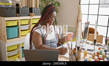 Beautiful african american woman artist painting a portrait, drawing on notebook whilst delightfully drinking morning coffee in her cozy art studio Stock Photo