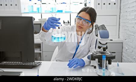 Hispanic woman scientist examining a flask with blue liquid in a modern laboratory setting. Stock Photo