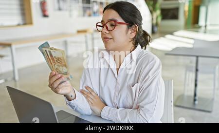 Young latina woman examines brazilian currency in a modern office setting, depicting business and financial themes. Stock Photo