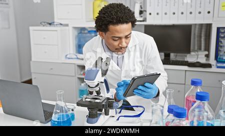 A focused man analyzing samples with a microscope and tablet in a modern laboratory. Stock Photo