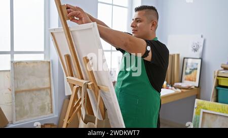 Young, confident latin artist joyfully standing by his easel in the art studio, ready to transfer creativity onto canvas Stock Photo