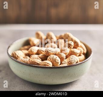 A bowl full of unshelled peanuts on a wooden table, suggesting a natural, healthy snack. Stock Photo