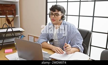 A young man wearing glasses works in an office, using a laptop and a headset while taking notes. Stock Photo