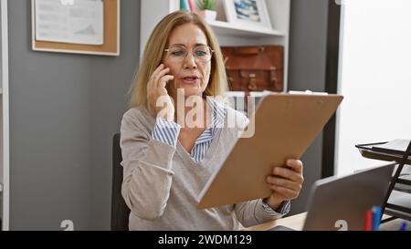 A professional woman converses on the phone while reviewing a document in her modern office. Stock Photo