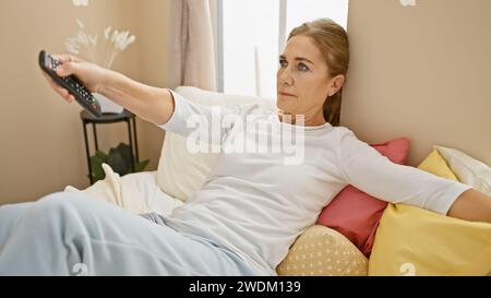 A mature woman relaxing at home, comfortably lounging on pillows while holding a remote control. Stock Photo