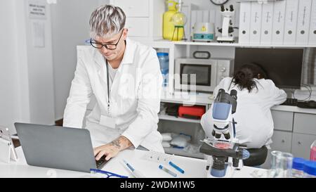 A man and woman working as coworkers in a laboratory, with him using a laptop and her examining samples under a microscope. Stock Photo