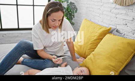 Loving mother measuring her sleepy daughter's temperature with a thermometer while she rests on the bed in their cozy bedroom Stock Photo