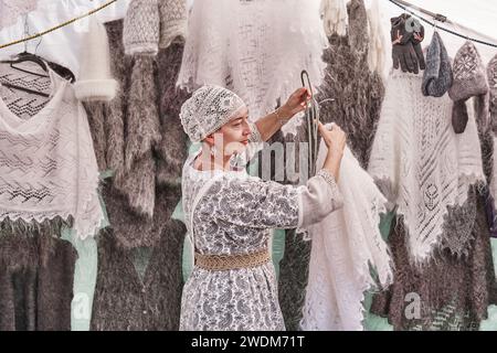 Senior caucasian woman hanging white fluffy shawl on hanger. She selling warm knitted items at fair. Stock Photo